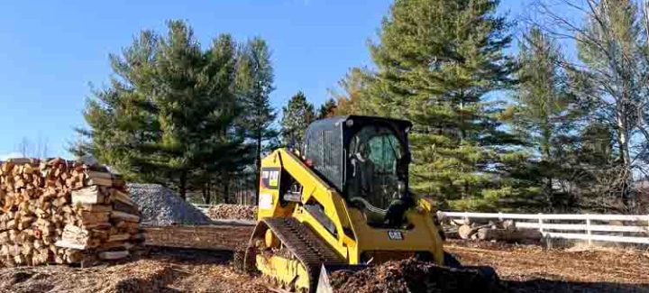 A compact yellow skid steer loader in a rural setting with a clear blue sky above. The machine is sitting on a bed of mulch, which could suggest recent landscaping or land-clearing work.
