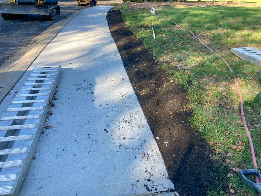 A freshly laid concrete pathway or sidewalk with a contrasting dark topsoil edge that has been neatly applied alongside it. This might be part of a landscaping effort to create a clean border between the walkway and the adjacent grassy area. On the left side of the image, there is a ladder lying flat, suggesting that work is either ongoing or has recently been completed in the area.